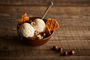 delicious ice cream with pieces of waffle, spoon and hazelnuts in bowl on wooden surface