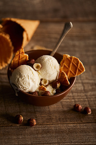 delicious ice cream with pieces of waffle, spoon and hazelnuts in bowl on wooden table