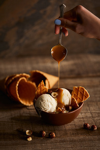 partial view of woman pouring caramel on delicious ice cream with pieces of waffle in bowl