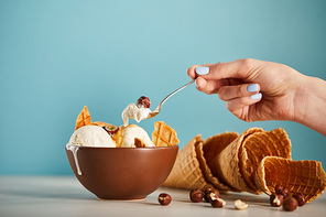 cropped view of woman with spoon, bowl of ice cream, hazelnuts and waffle cones on blue