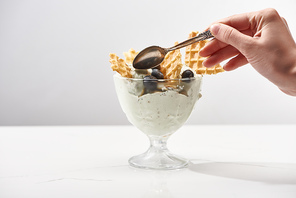 cropped view of woman holding spoon near bowl with pistachio ice cream with waffles and blueberries isolated on grey