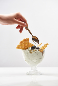 partial view of woman holding spoon near delicious pistachio ice cream with waffles and blueberries isolated on grey