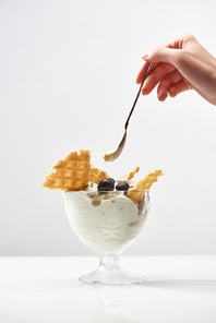 cropped view of woman holding spoon near delicious pistachio ice cream with waffles and blueberries isolated on grey