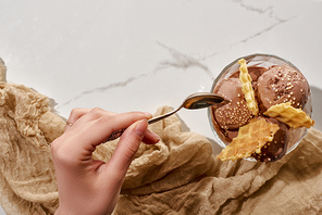cropped view of woman holding soon near delicious chocolate ice cream in bowl with waffles and brown cloth on marble grey background