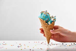 cropped view of woman holding delicious sweet blue ice cream with marshmallows and_sprinkles in crispy waffle cone isolated on grey