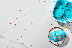 top view of delicious blue ice cream with sprinkles on coconut half and ice cream spoon on marble grey background