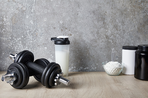 protein shake in sports bottle near dumbbells and protein powder near concrete wall