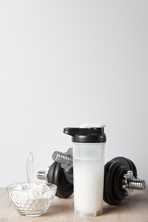 sports bottle with protein shake near black dumbbells and glass bowl with protein shake isolated on grey