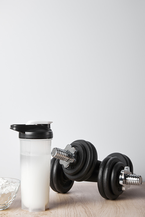sports bottle with protein shake near dumbbells isolated on grey