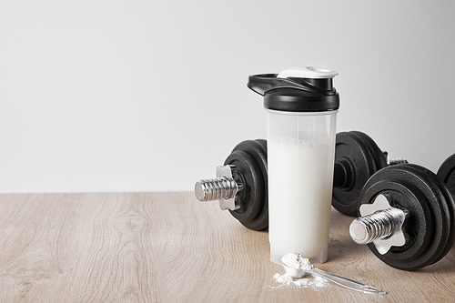 spoon with protein powder near heavy dumbbells and sports bottle isolated on grey