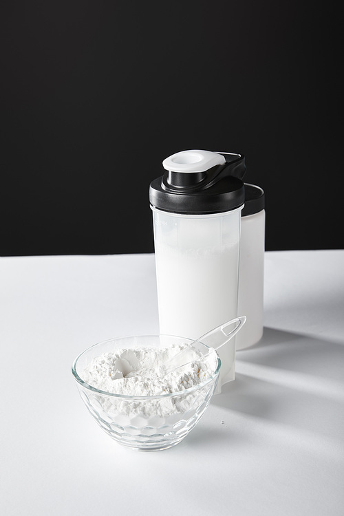 glass bowl with protein powder near sports bottle and jar on black