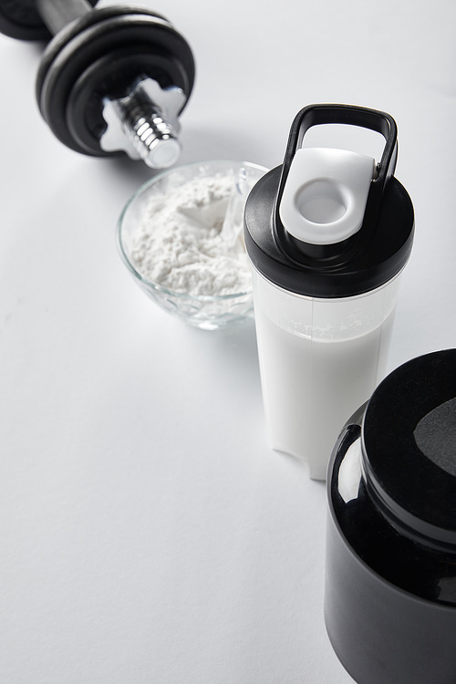 selective focus of sports bottle with protein shake near jar and dumbbell on white