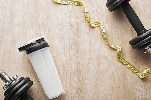 top view of dumbbells near sports bottle with protein shake near measuring tape on wooden surface