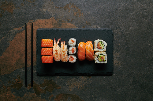 top view of arranged sushi set on black slate plate and chopsticks on dark surface