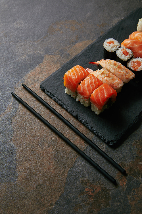 close up view of arranged sushi set on black slate plate and chopsticks on dark surface