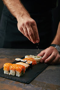partial view of chef pouring sesame seeds on arranged sushi set on slate plate on dark tabletop