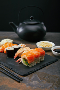 food composition with sushi set| ginger and soya sauce in bowls| teapot and cups with tea on dark surface