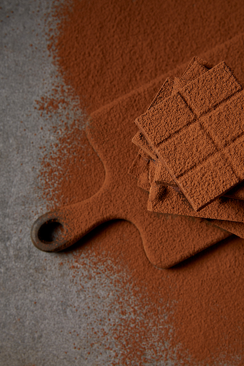 top view of sweet tasty chocolate pieces with cocoa powder on chopping board on grey