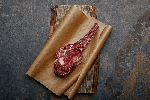 top view of raw rib eye steak on baking paper and wooden board on grey background