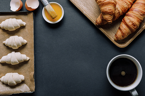 top view of dough for croissants on tray| egg yolk with brush and coffee cup on black surface