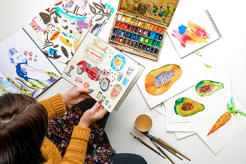 top view of woman holding album surrounded by color drawings and drawing utensils