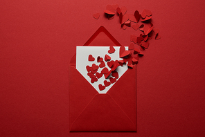 top view of white letter in envelope with pile of paper cut hearts on red background