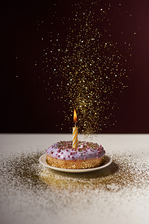 burning candle in middle of doughnut with falling sparkles on white table isolated on black