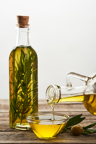 pouring olive oil from bottle into glass bowl| bottle of oil flavored with rosemary| olive tree leaves and olives on wooden surface