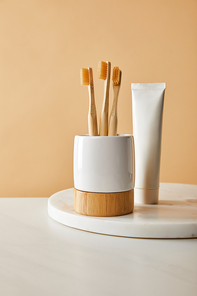 toothpaste in tube and holder with bamboo toothbrushes on white marble surface and beige background