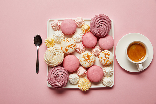 Flat lay with assorted meringues and macaroons on square dish with cup of coffee and spoon on pink background