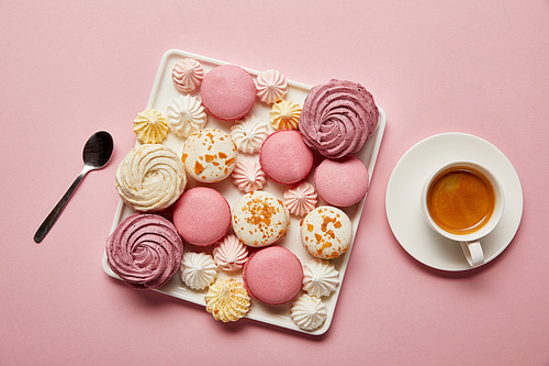 Top view of  meringues and macaroons on square dish with spoon and cup of tea on pink background