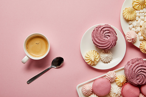 Top view of pink, white and yellow meringues, macaroons, marshmallows and cup of coffee with spoon on pink background