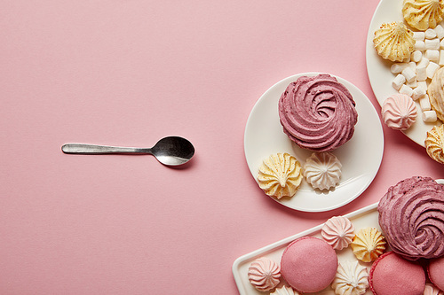 Plates with sweet meringues and macaroons with spoon on pink background