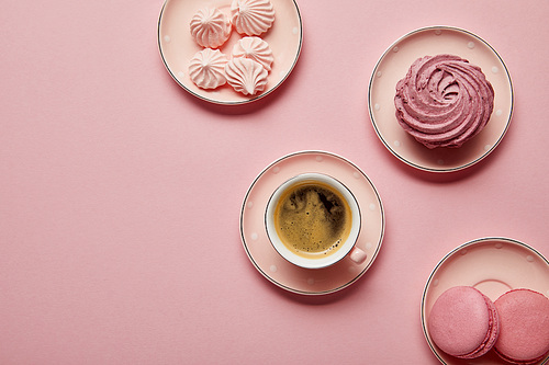 Top view of pink dotted saucers with meringues, macaroons and cup of coffee on pink background