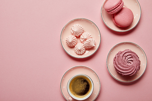 Top view of pink meringues and macaroons on small pink saucers with white dots and cup of coffee on pink background