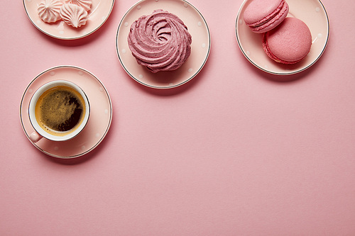 Top view of pink meringues, macaroons and cup of coffee on pink saucers with white dots on pink background