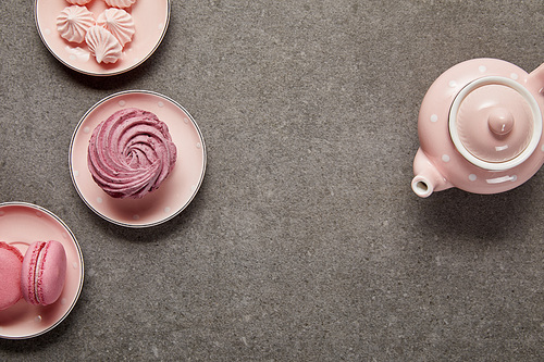 Top view of pink with dotted teapot and saucers with meringues and zephyr on gray background