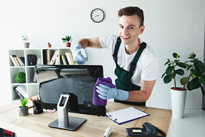 handsome young cleaner in rubber gloves cleaning computer monitor and smiling at camera