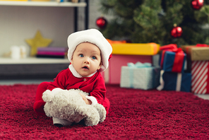 close-up portrait of adorable little baby in santa suit lying on red carpet with teddy bear in front of christmas tree and gifts