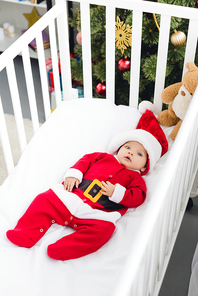 high angle view of beautiful little baby in santa suit lying in crib with teddy bear and 
