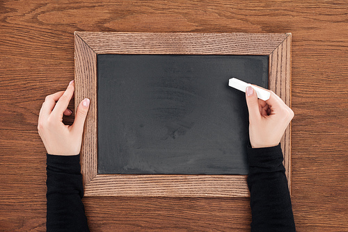 partial view of woman holding piece of chalk over empty chalk board on wooden background