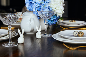 selective focus of decorative rabbits near plates, crystal glass and flowers on wooden table at home