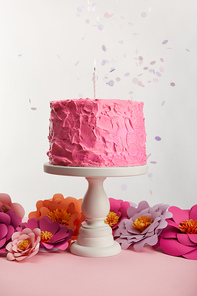 delicious pink birthday cake with candle on cake stand near paper flowers and confetti on grey