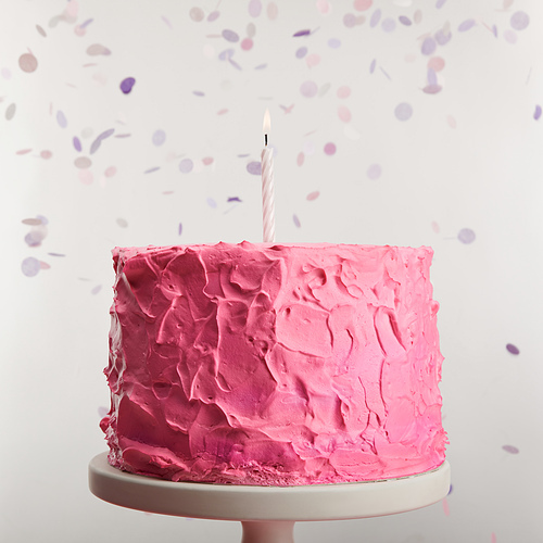 close up of delicious pink birthday cake with candle on cake stand near confetti on grey