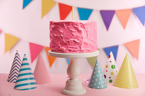 selective focus of delicious pink birthday cake with candle on cake stand near party caps and decoration