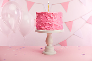 tasty pink birthday cake with burning candle on cake stand near air balloons and decoration