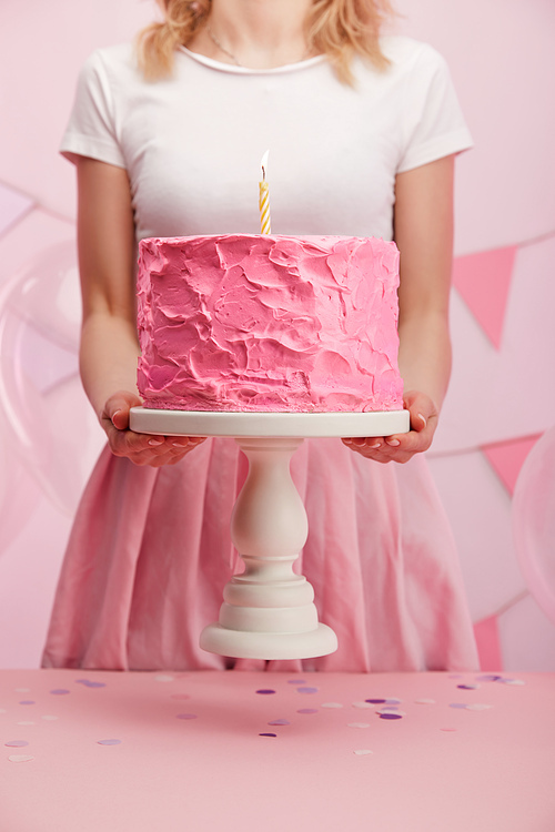 cropped view of woman holding cake stand with sweet pink birthday cake and burning candle