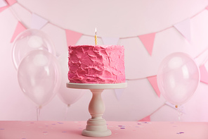 pink tasty birthday cake with burning candle on cake stand near air balloons and decoration