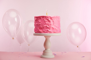 sweet and tasty birthday cake with burning candle on cake stand near air balloons on pink