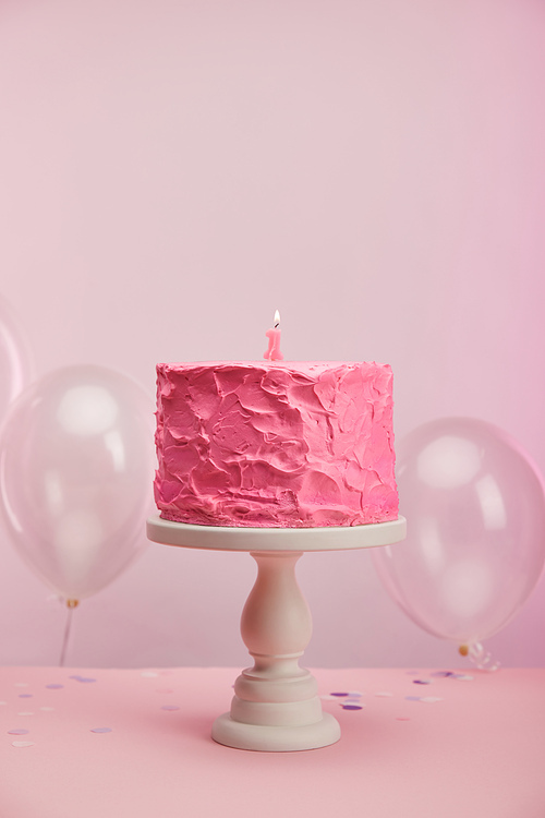 burning number one candle on pink and tasty birthday cake on cake stand near air balloons on pink